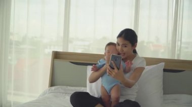  Genç Asyalı anne tatlı bir kız çocuğu tutuyor ve akıllı telefonuna el sallayarak konuşuyor. Yeni doğmuş tatlı bir kız bebek ağlıyor. konsept annelik, ebeveynlik çocuk odası aşkı ve aile hayatı