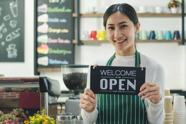 Çekici Asyalı kadınlar kahvede gülümsüyor ve kendilerine güveniyorlar. Genç bayan barista, kafeterya sahibi, açık tabela tutuyor ve barın girişine hoş geldiniz. Restoranlarda garsonluk ve önlük giyme işi