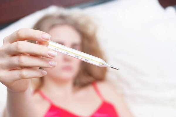 Женщина с вирусом гриппа лежит в постели и измеряет температуру термометром.
