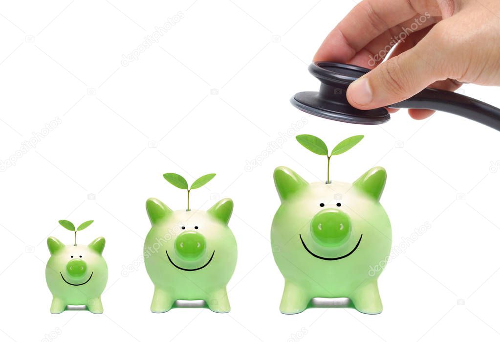 Hand holding a stethoscope over green piggy banks with young green plants isolated on white      
