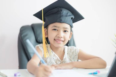 Küçük Asyalı kız ödev ve gülümseme yapıyor lisansüstü şapka giyiyor 