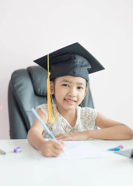 Маленькая азиатка в шляпе выпускника делает домашнее задание и улыбается — стоковое фото