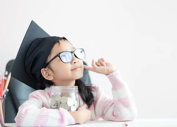 Little Asian girl wearing graduate hat hugging clear glass jar p — Stock Photo, Image