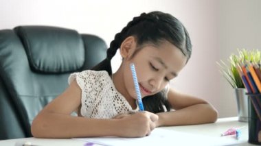 Küçük Asyalı kız ödevlerini yapmak için kalemi kullanıyor ve eğitim konsepti için mutlulukla gülümsüyor.