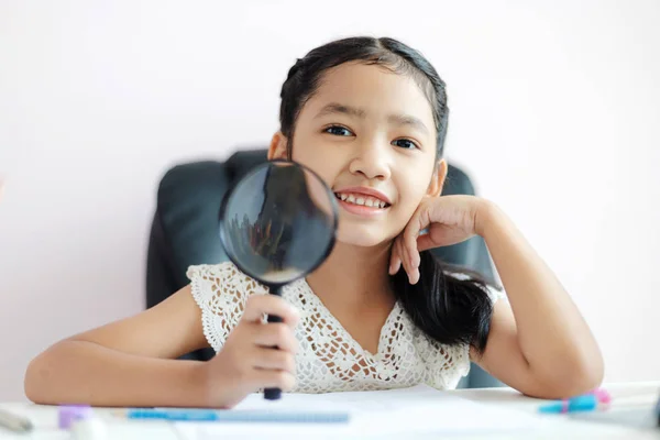 Liten asiatisk tjej med förstoringsglas gör läxor och Le med — Stockfoto