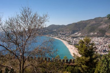 Ağaçlar ve mavi denizin üzerinde yükselen bir kale duvarı.