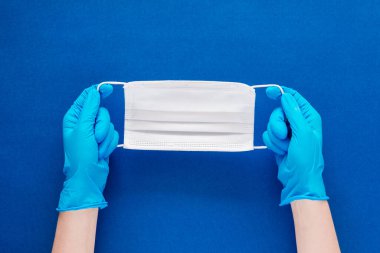 Mavi eldivenli, mavi desenli bir arka planda maske tutan bir kadının el resmi.