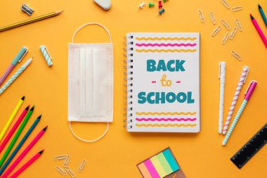 Okul konseptinin yeni normal haliyle çekilmiş resmi maske takılı bir defter ve sarı arka planda birkaç kırtasiye eşyası.