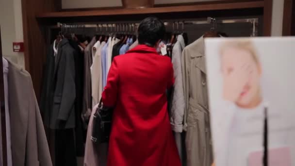 Женщина в красном платье выбирает одежду в магазине одежды . — стоковое видео