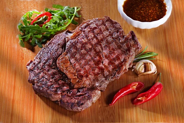 Juicy delicious medium fried meat steak on a wooden cutting board, with pepper, garlic, herbs and tomato sauce. Steak for a restaurant on wooden background — Stock Photo, Image