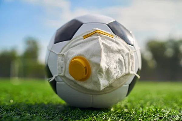 Medikal Maskeli Futbol Balosu Stadyumun Çimlerinde Yatar. Futbol topunda vanası olan bir solunum cihazı. Covid-19 'dan koruma                   