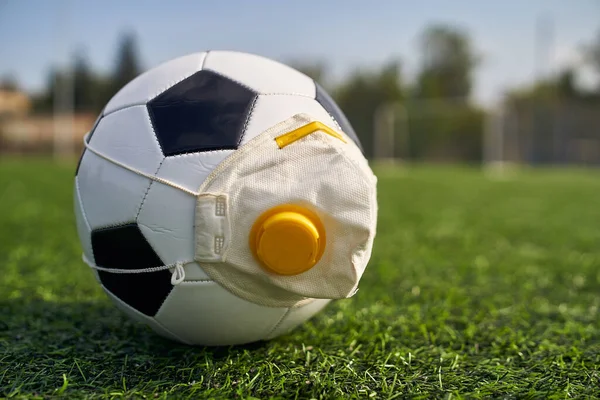 Medikal Maskeli Futbol Balosu Stadyumun Çimlerinde Yatar. Futbol topunda vanası olan bir solunum cihazı. Covid-19 'dan koruma                   