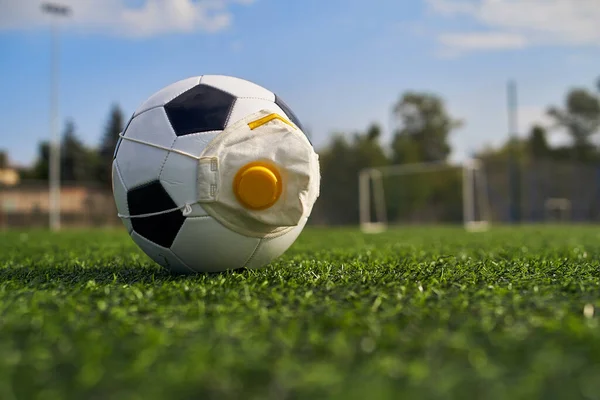 Medikal Maskeli Futbol Balosu Stadyumun Çimlerinde Yatar. Futbol topunda vanası olan bir solunum cihazı. Covid-19 'dan koruma                   