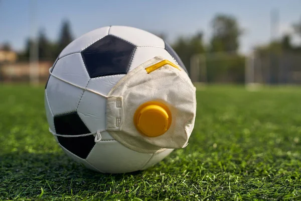 Medikal Maskeli Futbol Balosu Stadyumun Çimlerinde Yatar. Futbol topunda vanası olan bir solunum cihazı. Covid-19 'dan koruma                   