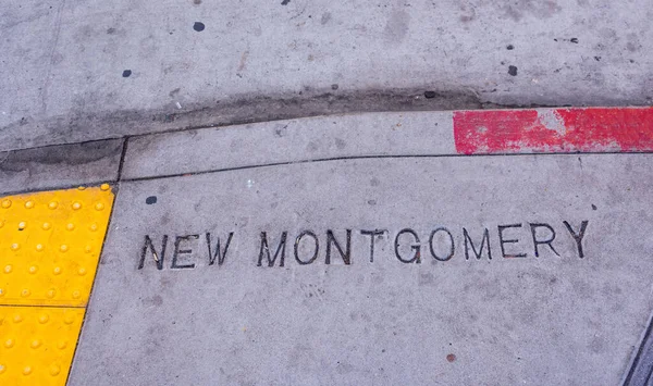New Montgomery street name sign stamped on a sidewalk in downtown San Francisco. Yellow ADA compliant tactile paving. Weathered red paint on curb. Little black gum dots on concrete sidewalk.
