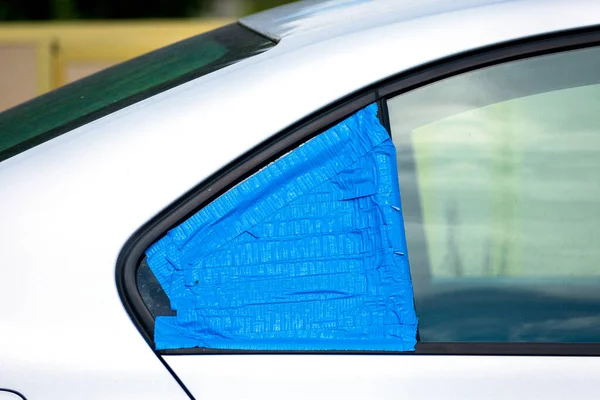 Broken and damaged car window temporarily covered with blue tape to protect interior from rain and water. Smash-and-grab automobile break-ins. Insurance concept.
