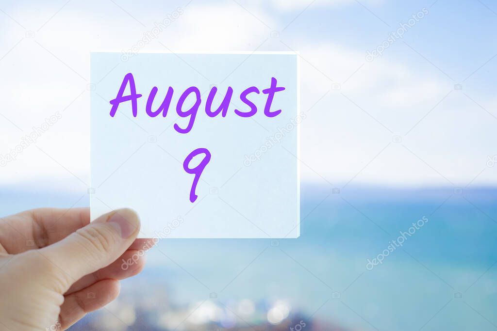 August 9th. Hand holding sticker with text August 9 on the blurred background of the sea and sky. Copy space for text. Month in calendar concept.