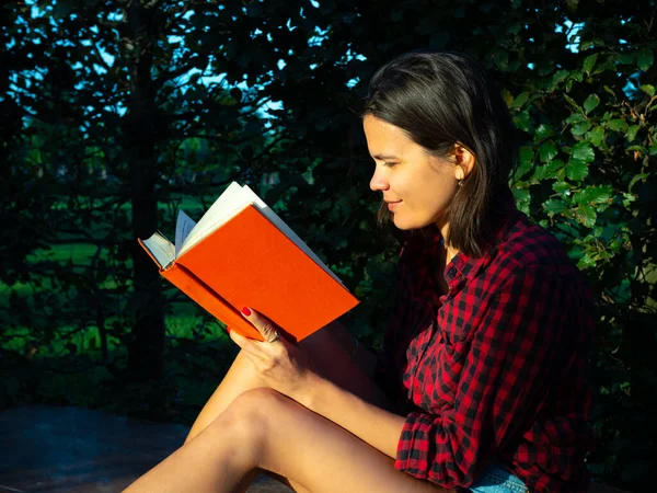 Giovane ragazza adulta caucasica in camicia rossa si siede nel parco al mattino e legge libro — Foto Stock
