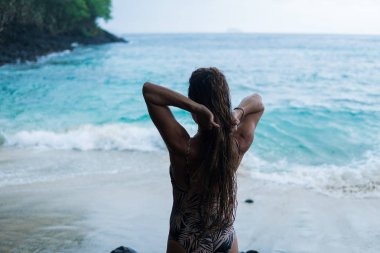 güzel uzun saçlı tabaklanmış kız Avrupa görünüm okyanus plaj, taş ve ağaç arka plan üzerinde kamera için poz. Endonezya, Bali