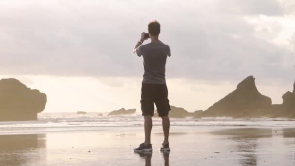 Άνθρωπος τουρίστας λήψη φωτογραφιών σε smartphone κάμερα στην παραλία στο φόντο του ωκεανού και βράχια. Μπαλί, Ινδονησία — Αρχείο Βίντεο
