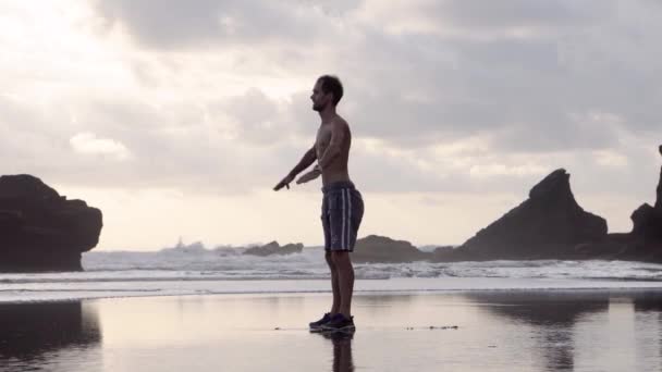 Young man in shorts and sneakers doing stretching exercises on a beach with rocks early in the morning or evening — Stockvideo