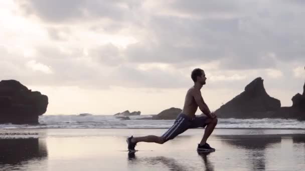 Νεαρός άνδρας με σορτς και αθλητικά κάνοντας ασκήσεις τεντώματος σε μια παραλία με βράχια νωρίς το πρωί ή το βράδυ — Αρχείο Βίντεο