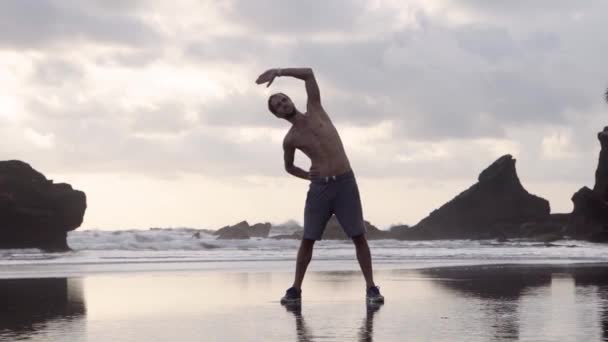 Joven en pantalones cortos y zapatillas de deporte haciendo ejercicios de estiramiento en una playa con rocas temprano en la mañana o por la noche — Vídeos de Stock