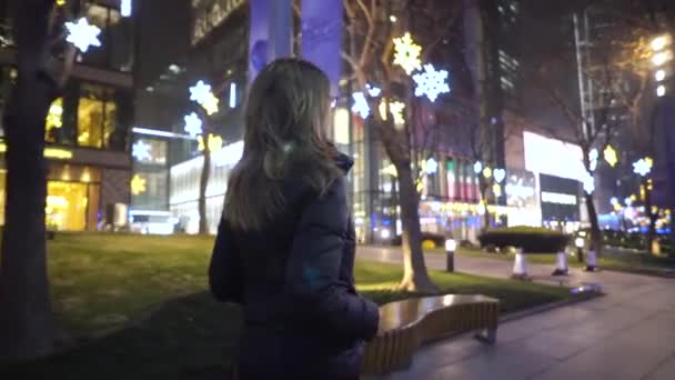 Una joven vestida con una chaqueta negra y gafas, pasea lentamente por el centro de la ciudad entre tiendas y rascacielos — Vídeos de Stock