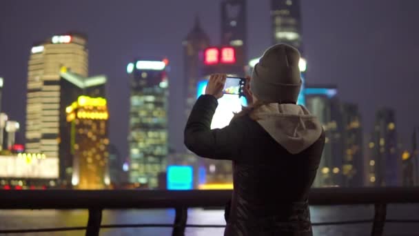 Una joven con una chaqueta negra y sombrero toma fotos de atracciones Shanghais en el teléfono móvil — Vídeos de Stock