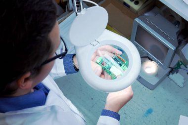 Beyaz cüppe li ve gözlüklü bir adam büyüteçle elektrik panosunu kontrol eder. Üretimde kalite kontrol departmanı. yüksek teknoloji fabrikasında üretim