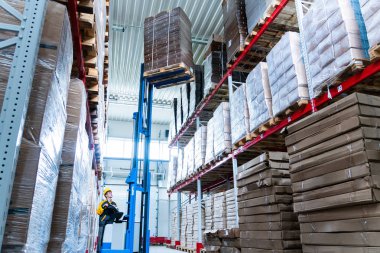 Üniformalı depo işçisi forklift yükleyicisiyle karton kutular yüklüyor.