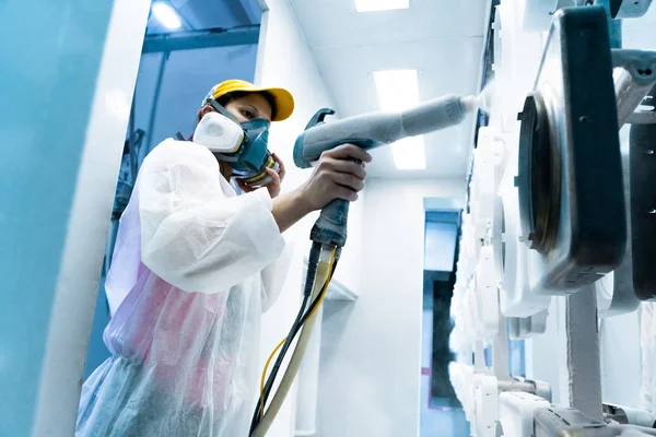 Powder coating of metal parts. A woman in a protective suit sprays white powder paint from a gun on metal products