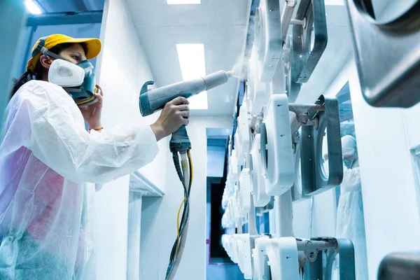 Revestimento em pó de peças metálicas. Uma mulher em um terno protetor pulveriza tinta branca em pó de uma arma em produtos metálicos — Fotografia de Stock