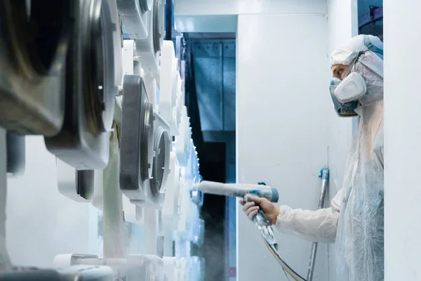 Recubrimiento en polvo de piezas metálicas. El hombre con traje protector rocía pintura en polvo blanca de una pistola sobre productos metálicos — Foto de Stock