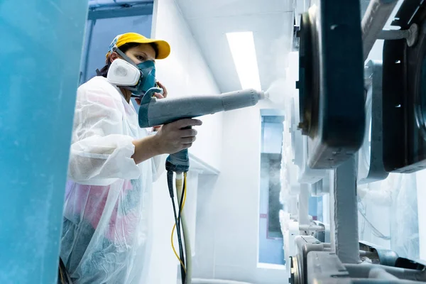 Powder coating of metal parts. A woman in a protective suit sprays white powder paint from a gun on metal products