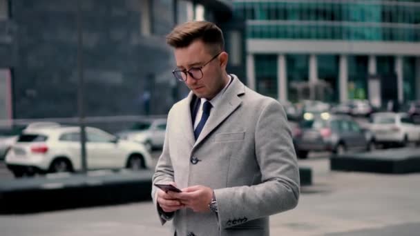 Un uomo d'affari con un cappotto grigio e occhiali usa uno smartphone, legge le notizie nel centro commerciale della città, tra i grattacieli — Video Stock