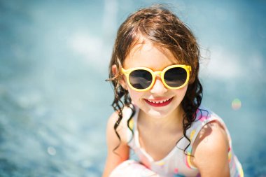Arka planda mavi su bulunan, yaz sarısı güneş gözlüklü, gülümseyen küçük kızın yakın plan portresi. Denizde yaz tatili. Sıcak yaz gününde mutlu bir çocuk. Metin için alanı kopyala.