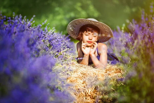 美丽的小女孩穿着蓝色的衣服 头戴白色的帽子 闭着眼睛躺在薰衣草地里 夏日阳光灿烂 周围开着紫罗兰的花朵 普罗旺斯的夏天旅行 薰衣草中的儿童 — 图库照片