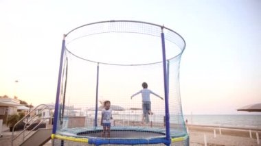 Gün batımında iki çocuk plajda trambolinde zıplıyor. Oğlan ve kız denizde eğlenceli bir yaz tatili geçiriyorlar. Çocukluk ve aktif yaşam tarzı kavramı.