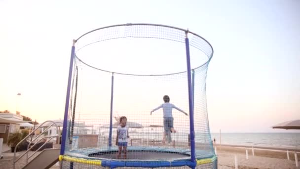 Dos niños saltan en un trampolín en la playa al atardecer. Chico y chica divertirse vacaciones de verano en el mar. Concepto de infancia y estilo de vida activo . — Vídeo de stock