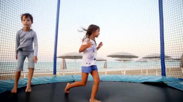 Felices hermanos sonriendo niños rebotando saltando en el trampolín en la playa durante el atardecer. Los niños tienen vacaciones divertidas en el mar. Feliz infancia y concepto de estilo de vida activo . — Vídeo de stock