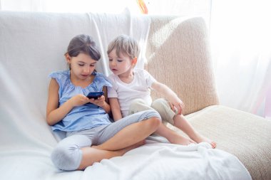 Şirin küçük kardeşler ev kavramında iyi vakit geçiriyorlar. Yakın küçük kardeş tabletle oynuyor. Çocuklar kanepede oturup tabletle oynuyorlar. Çocuklar cep telefonuyla oyun oynuyorlar.