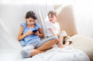 Çocuklar kanepede oturup tabletle oynuyorlar. Sevimli erkek ve kız kardeşler evde iyi vakit geçiriyorlar. Çocuklar cep telefonuyla oyun oynuyorlar. COVID-19 konsepti süresince evde kalın