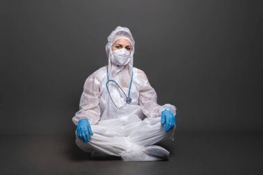Doktorlar Covid-19 salgını sırasında çalışırlar. Koruyucu takım elbiseli ve maskeli yorgun kadın doktor gri arka planda yerde oturuyor..