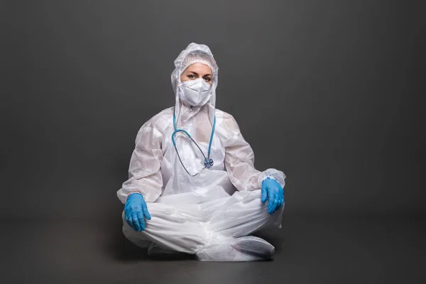 Doctors work during Covid-19 pandemic concept. Tired female doctor in a protective suit and mask sitting on the floor on a gray background.