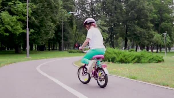 Το χαριτωμένο κοριτσάκι αρχίζει να κάνει ποδήλατο στο πάρκο. Ένα κορίτσι με προστατευτικό κράνος. Έννοια της ασφάλειας των παιδικών δραστηριοτήτων. — Αρχείο Βίντεο