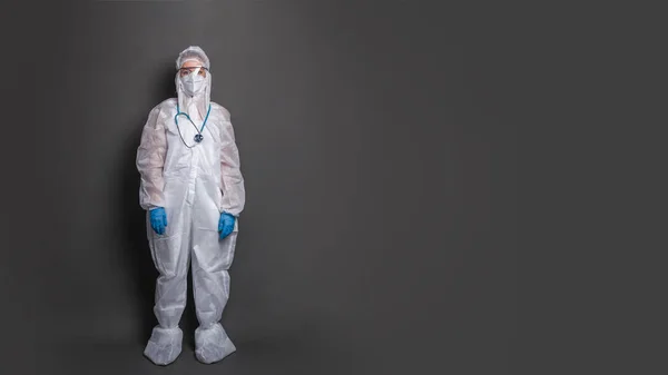 Doctor wearing PPE and working during the coronavirus pandemic concept. A young female doctor in a protective suit stands in full growth on a gray background.