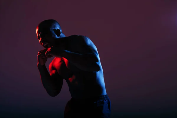 An African American in a red-blue neon color with a naked torso stood in a combat pose in the studio. Advertising sports shooting
