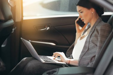 Takım elbiseli genç bir iş kadını arabada otururken yatırımcılarla konuşuyor ve laptoptan bilgileri kontrol ediyor..
