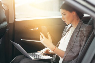 Takım elbiseli güzel bir kadın yolcu kompartımanında oturuyor ve akıllı telefon ve dizüstü bilgisayarla verileri kontrol ediyor.